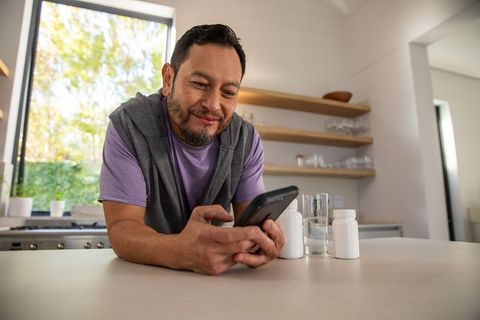 Mature asian man checking smartphone while leaning on modern kitchen counter with meds