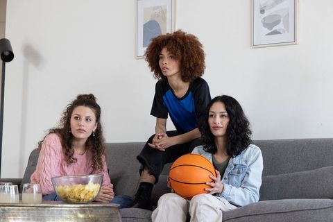 Diverse young women on couch watching game, sharing snacks and holding basketball