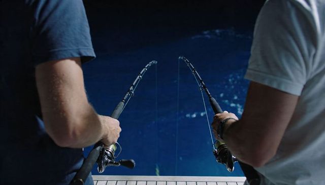 Two anglers night fishing on boat deck holding spinning rods over deep blue ocean