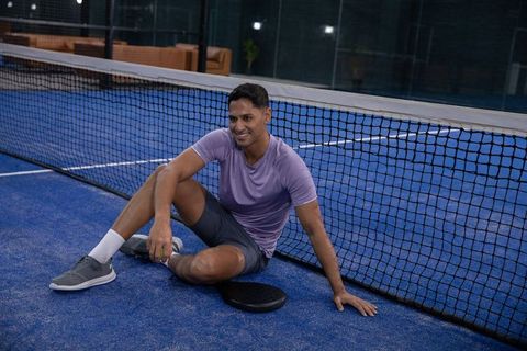 Smiling man resting on indoor padel court holding green ball by net and racket