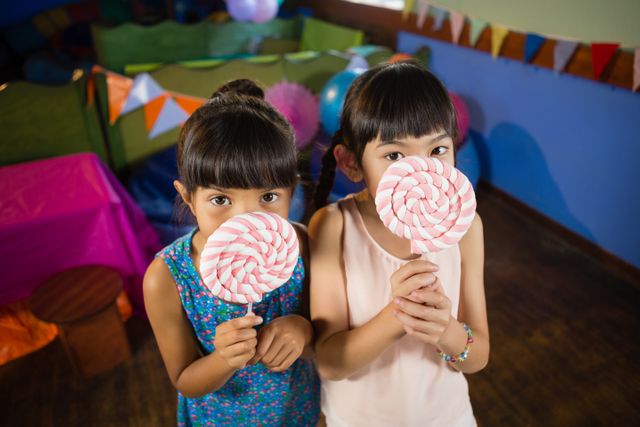 Two children holding large lollipops, standing indoors at a birthday party. The scene is colorful and festive, with decorations in the background. Ideal for use in content related to children's parties, celebrations, sweets, and childhood fun.