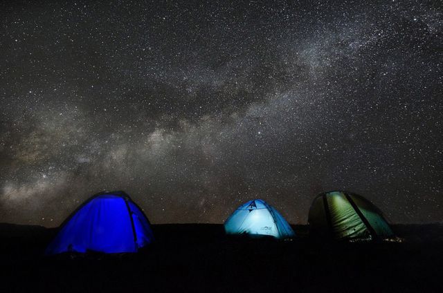 Three illuminated tents on dark ground beneath a night sky full of stars and the Milky Way visible in the background. Ideal for use in travel blogs, outdoor adventure posters, astronomy websites, stargazing guides, nature enthusiasts marketing materials.