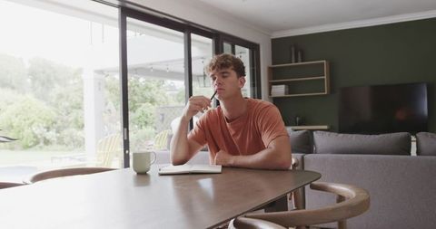 Young man contemplating while writing in notebook at sunlit modern dining table with patio