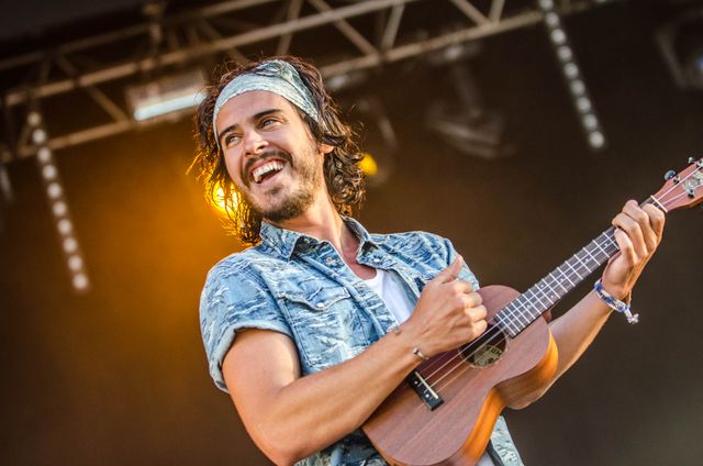 Male musician playing ukulele and smiling energetically on stage during a live concert. The performer, dressed casually in a jean shirt and headband, captivates the audience with his cheerful demeanor under the spotlights at an outdoor summer festival. Ideal for use in promoting live music events, summer festivals, and in articles about the joy of live performances.