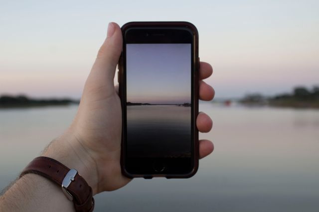 Hand holding smartphone showing a tranquil lake scene during twilight with calm waters reflecting soft sky hues. Ideal for concepts of mobile photography, technology in nature, scenic captures, or social media content production.