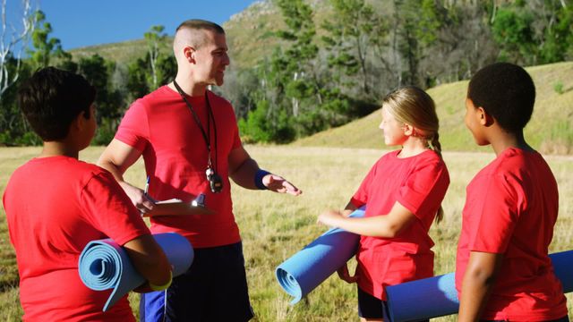 Male trainer wearing red shirt interacting with kids holding yoga mats on sunny day. Diverse group of children attentively listening. Suitable for depicting fitness training, youth mentorship, teamwork in summer camps, and educational outdoor activities. Ideal for promotional materials related to camps, fitness programs for kids, health awareness initiatives, team-building exercises, and community engagement programs.