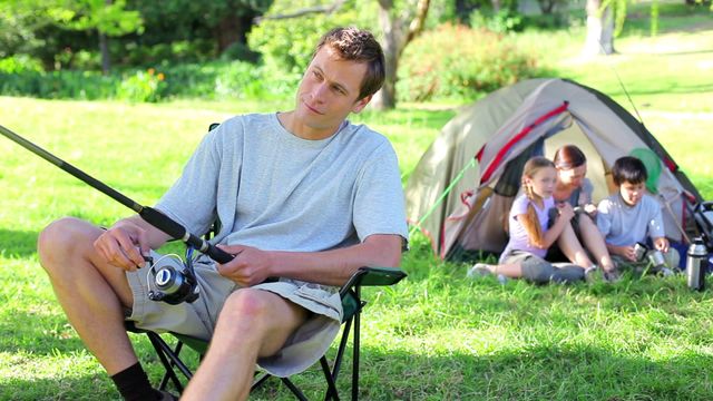 The scene shows a father enjoying fishing while camping with his family in the countryside. A tent is pitched in the background with children relaxing on the grass alongside another family member. This image is ideal for promoting family bonding activities, outdoor adventures, camping gear, and leisure time spent in nature.