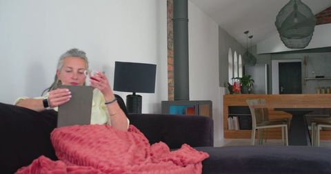 Relaxing woman using tablet and holding cup under pink throw in modern open-plan living room