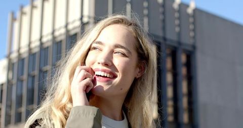 Sunlit Blonde Woman Smiling While Talking on Smartphone Outside Modern Office Building