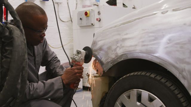 Skilled auto mechanic expertly uses angle grinder on car panel to repair damage in bustling workshop environment. Sparks from grinder show focus and intense precision of mechanics. Ideal for showcasing craftsmanship, automotive skills, or industrial maintenance themes.