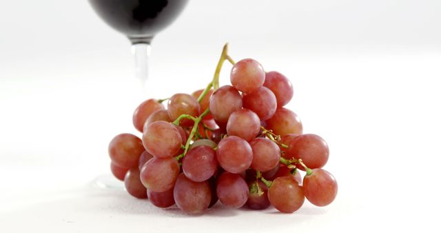 Close-up shot of a bunch of red grapes with a glass of red wine in the background on a white surface. Perfect for use in food and beverage advertisements, healthy eating and lifestyle promotions, recipe blogs, and as a decorative piece for restaurants and vineyards.
