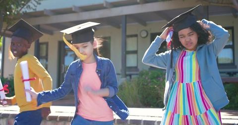 Diverse children celebrating graduation walking along brick wall holding diplomas