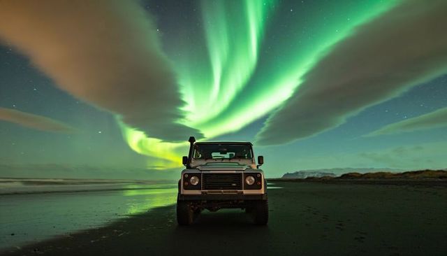 Boxy 4x4 parking on black sand beach under vivid aurora borealis and sky reflections