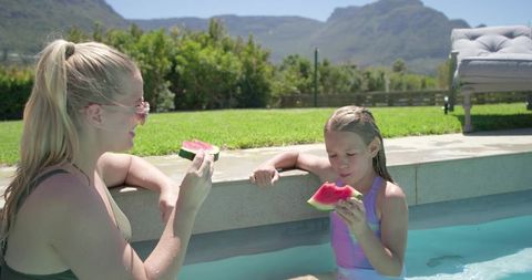 Woman and girl sharing watermelon by backyard pool, sunlit summer with mountain view