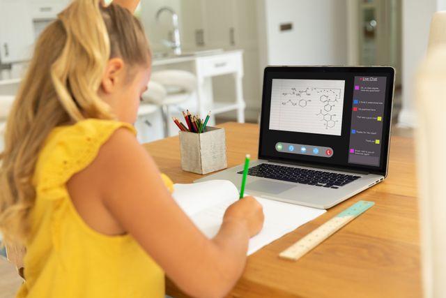 Young girl participates in online lecture on laptop, writing notes in a notebook. Useful for content focused on e-learning, remote education, child development, homeschooling, and modern technology in education.