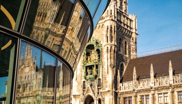 Curved glass facade reflecting ornate gothic town hall with clock figurines in urban plaza