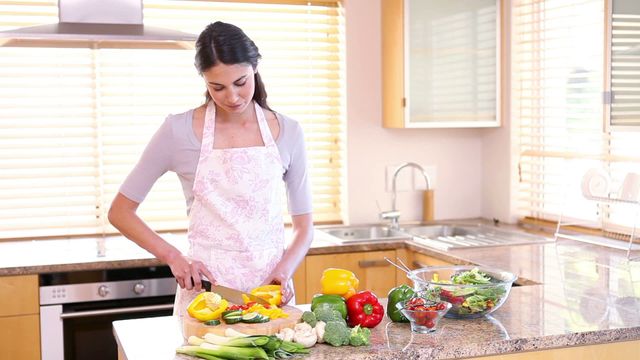 A woman wearing an apron cuts fresh vegetables in a bright kitchen. The scene includes various vegetables on a wooden counter, highlighting healthy cooking and meal preparation. Ideal for content focusing on healthy eating habits, home cooking, kitchen design, culinary blogs, or advertisements promoting kitchenware or healthy living.