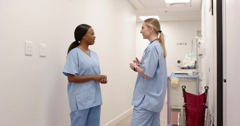 Two nurses talking in bright hospital corridor with wheelchair and medical equipment