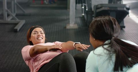 Diverse women doing partner sit-ups in gym wearing smartwatch and athleticwear, smiling