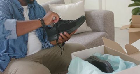 African american man unboxing black sneakers and testing laces on couch in living room