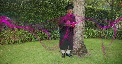 Graduating student standing by tree in gown and mortarboard with magenta wave overlay, garden backdr