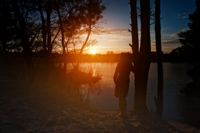 Silhouette of a woman standing by a lake, gazing at the sunset. Ideal for themes of tranquility, solitude, and nature's beauty. Could be used for environmental awareness campaigns, meditative or inspirational content, and travel advertisements highlighting serene destinations.