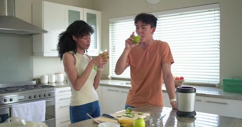 Multicultural couple making green smoothies at modern kitchen island with blender and fruit