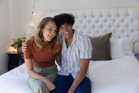 Smiling young couple sitting on bed leaning together by tufted headboard with cozy string lights