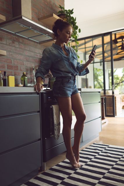 Woman standing in a modern kitchen, using a mobile phone. She is dressed in casual denim clothing and appears relaxed. This image can be used for lifestyle blogs, technology advertisements, home decor magazines, or articles about modern living and communication.