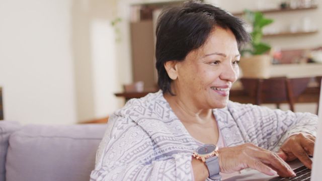 Video depicts a senior African American woman using a laptop while sitting on a couch at home. She is smiling and appears to be enjoying her time, signifying a relaxed and comfortable lifestyle during retirement. This can be used for concepts related to technology usage among elderly, retirement living, happiness in later life, or showcasing independence in seniors.