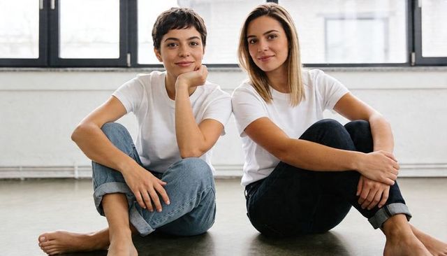 Two women sitting on studio floor wearing white tees and jeans, casual friendship portrait