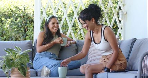Two friends relaxing on patio sofa sipping coffee and laughing in sunny outdoor living space