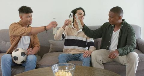 Multicultural friends clinking bottles on couch while watching soccer and sharing snacks