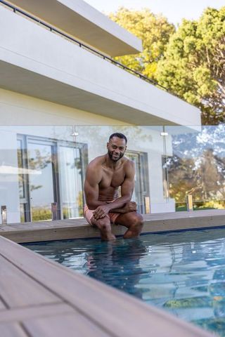 African american man sitting on modern pool deck, relaxing with feet in water, summer lifestyle