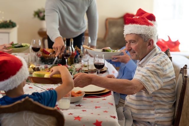 Elderly man wearing Santa hat helping his grandson with meal at Christmas dinner table. Family members enjoying festive meal together, creating a warm and joyful atmosphere. Ideal for use in holiday greeting cards, family-oriented advertisements, or articles about holiday traditions and family bonding.