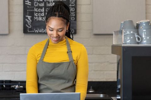 Smiling african american barista typing on laptop at cafe counter with espresso machine