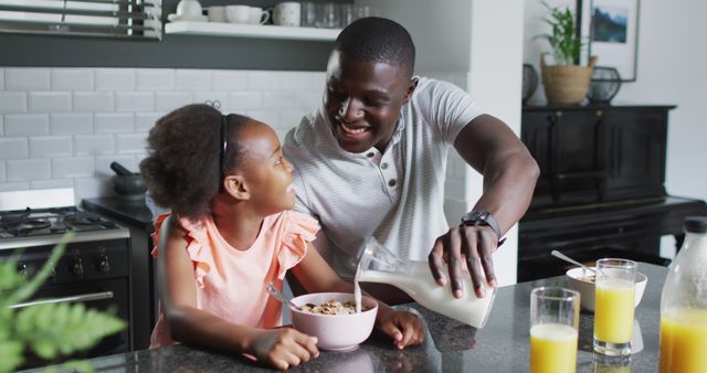 Father pouring milk into daughter's cereal while both are smiling in a modern kitchen. Perfect for themes related to parenting, family bonding, morning routines, nutrition, or modern lifestyles. Can be used in articles, blogs, or advertisements promoting healthy family habits, nutritious breakfasts, or quality family time.