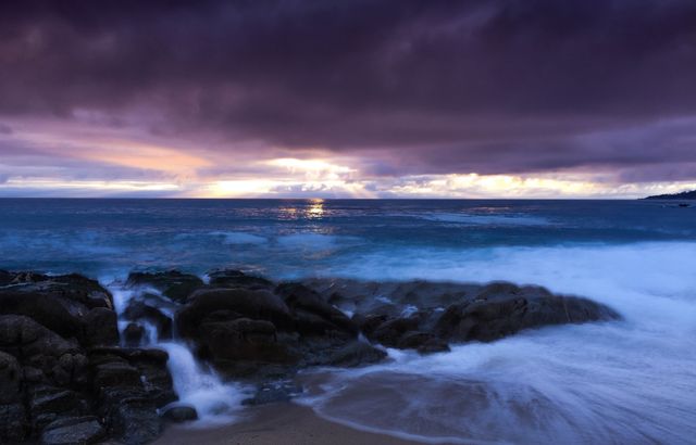 This image captures a dramatic sunset over a rocky seashore with purple clouds filling the sky. It showcases the natural beauty of the ocean waves crashing against rocks under the twilight. Ideal for use in travel brochures, nature magazines, desktop wallpapers, or peaceful retreat promotions.