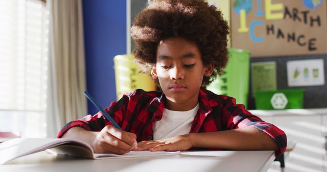 Young African American boy wearing red and black checkered shirt writing attentively at desk in classroom with educational posters in background. Useful for educational content, school brochures, or articles about childhood learning and student engagement.