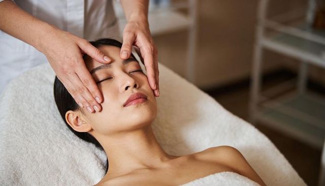 Asian woman receiving relaxing facial massage on spa bed with towel, eyes closed