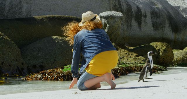 The scene captures a woman kneeling on a sandy beach, reaching towards a nearby penguin close to the water. She is wearing a denim jacket, yellow dress, and straw hat. Use this for travel themes, wildlife interactions, or scenes of nature and leisure activities near the ocean.