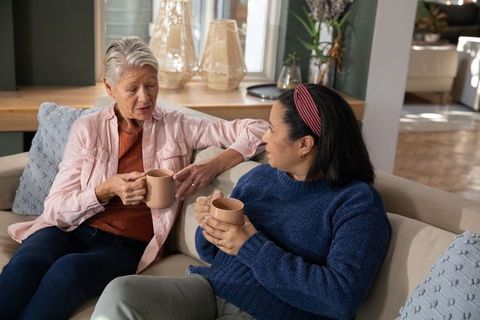 Senior friends sharing coffee on sofa, cozy conversation in sunlit living room