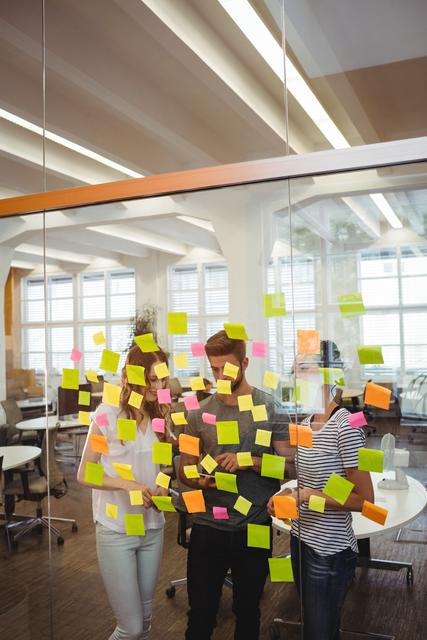 Three business executives are collaborating in an office, using sticky notes on a glass wall for brainstorming and planning. They are discussing ideas while looking at a digital tablet. This image can be used for depicting teamwork, creativity, and modern professional environments in presentations, articles, and websites.