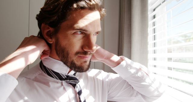 Young man adjusting necktie in front of window with natural light streaming through blinds. Perfect for illustrations depicting daily routines, preparation for professional events, or advertisements focused on men's fashion and grooming.