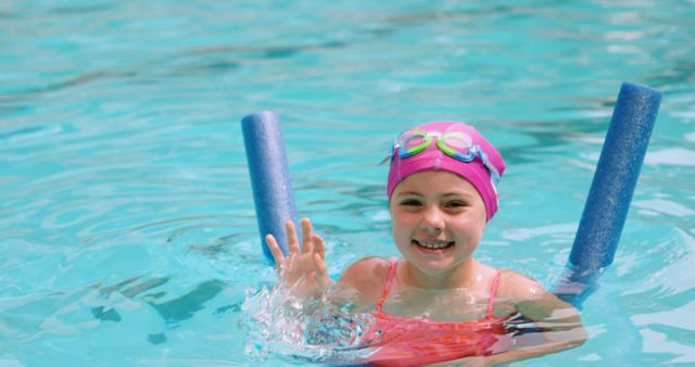 Young girl swimming in pool, wearing pink swim cap and goggles, holding blue pool noodles. She is smiling and waving, indicating enjoyment and confidence in water. Ideal for use in content related to summer activities, children's swimming lessons, water safety, and outdoor fun.