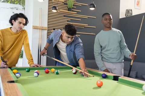 Diverse male friends playing pool in modern lounge room, teamwork and leisure