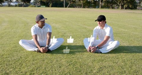 Two Teammates Sitting on Grass Field Smiling and Stretching in White Polo Cricket Attire