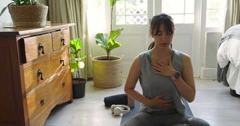Asian woman practicing breathing exercise on yoga mat in sunlit room