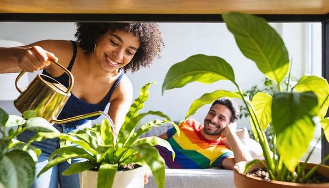 Smiling woman watering indoor plants while partner relaxing on sunlit living room couch