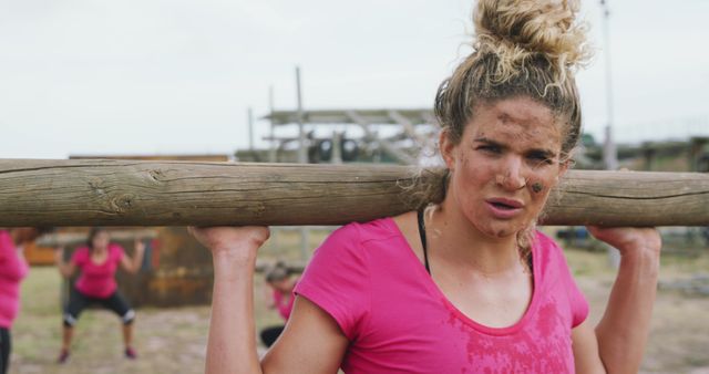 Woman wearing pink shirt involved in outdoor obstacle course carrying heavy log on her shoulders. Face shows determination and is slightly muddy, highlighting the intensity of the workout. Ideal for fitness, perseverance, and team-building concepts, this image is perfect for advertisements, promotional materials, and articles on outdoor fitness challenges and training programs.